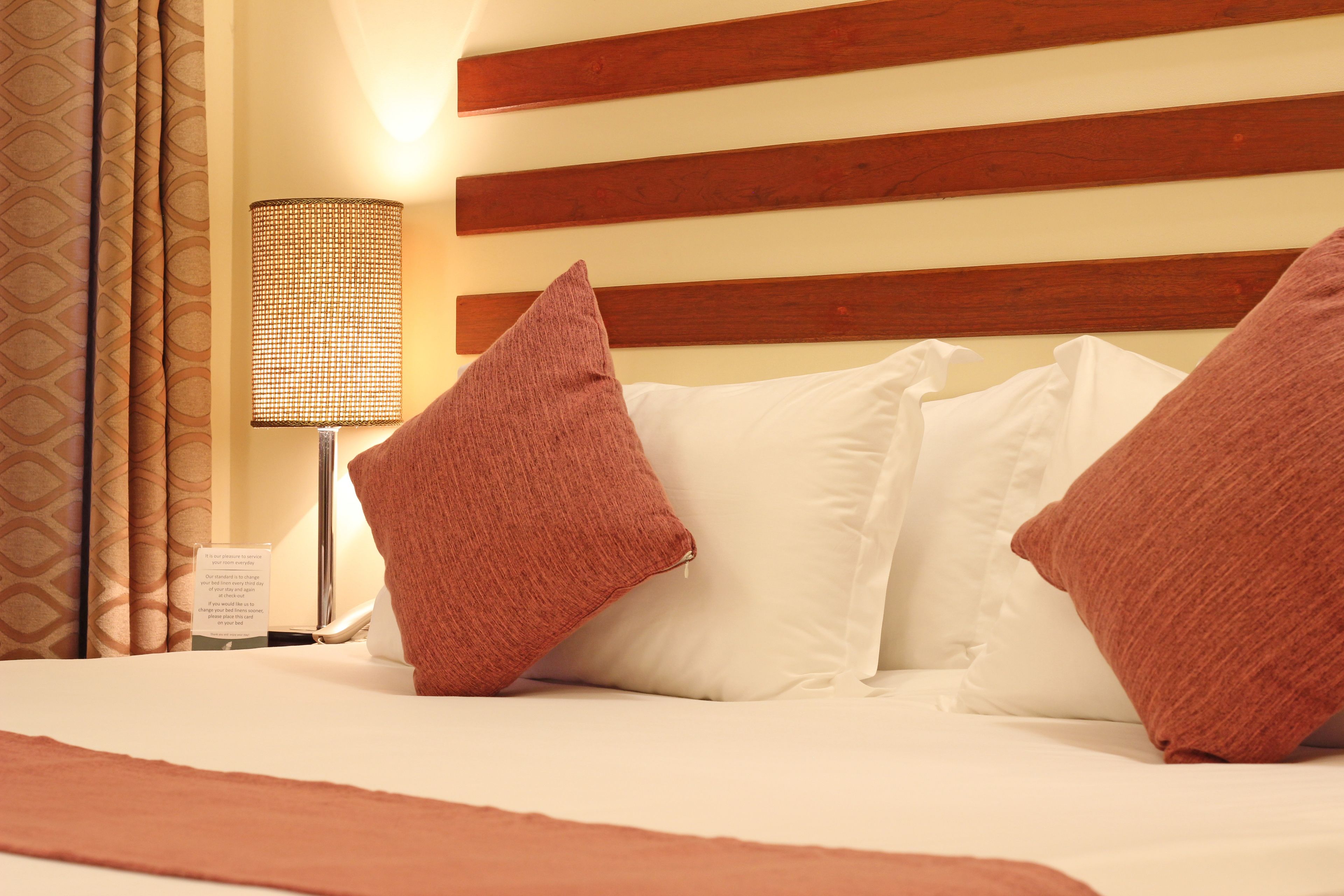 A hotel room with a bed, pillows, a lamp, and a sign on the nightstand.