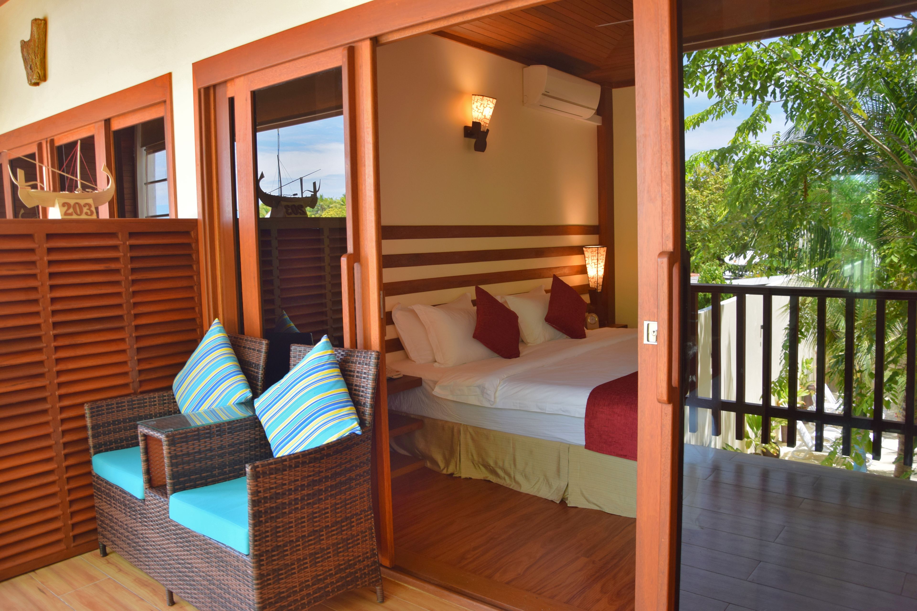 A room with a bed, a wicker chair with cushions, a wooden ceiling, and a view of greenery outside.