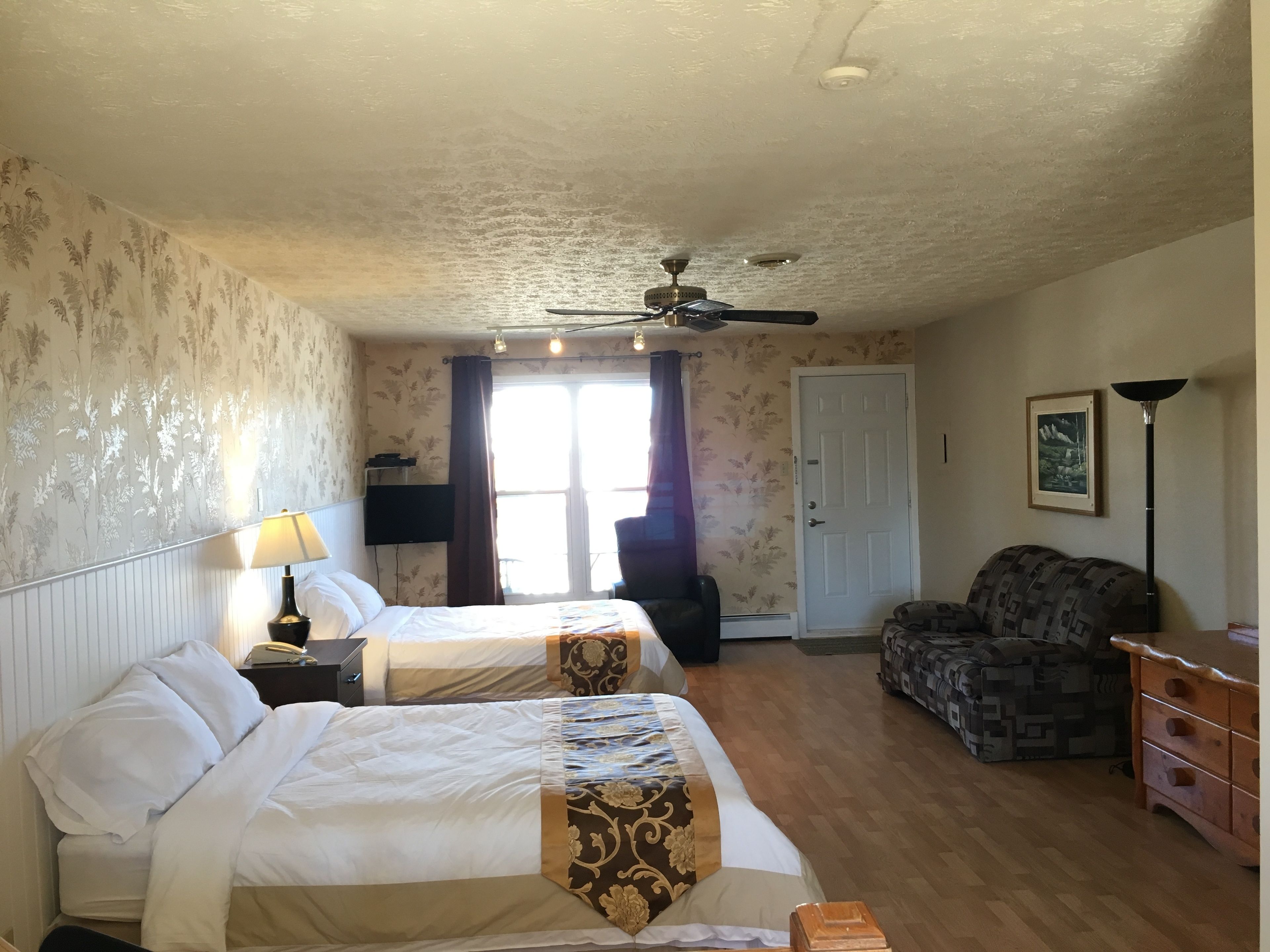 A hotel room with two beds, a ceiling fan, a window with curtains, a sofa, a lamp, and a framed picture on the wall.