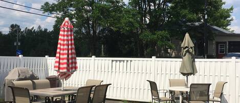 An outdoor patio with a table and chairs, a red and white striped umbrella, and a white picket fence.