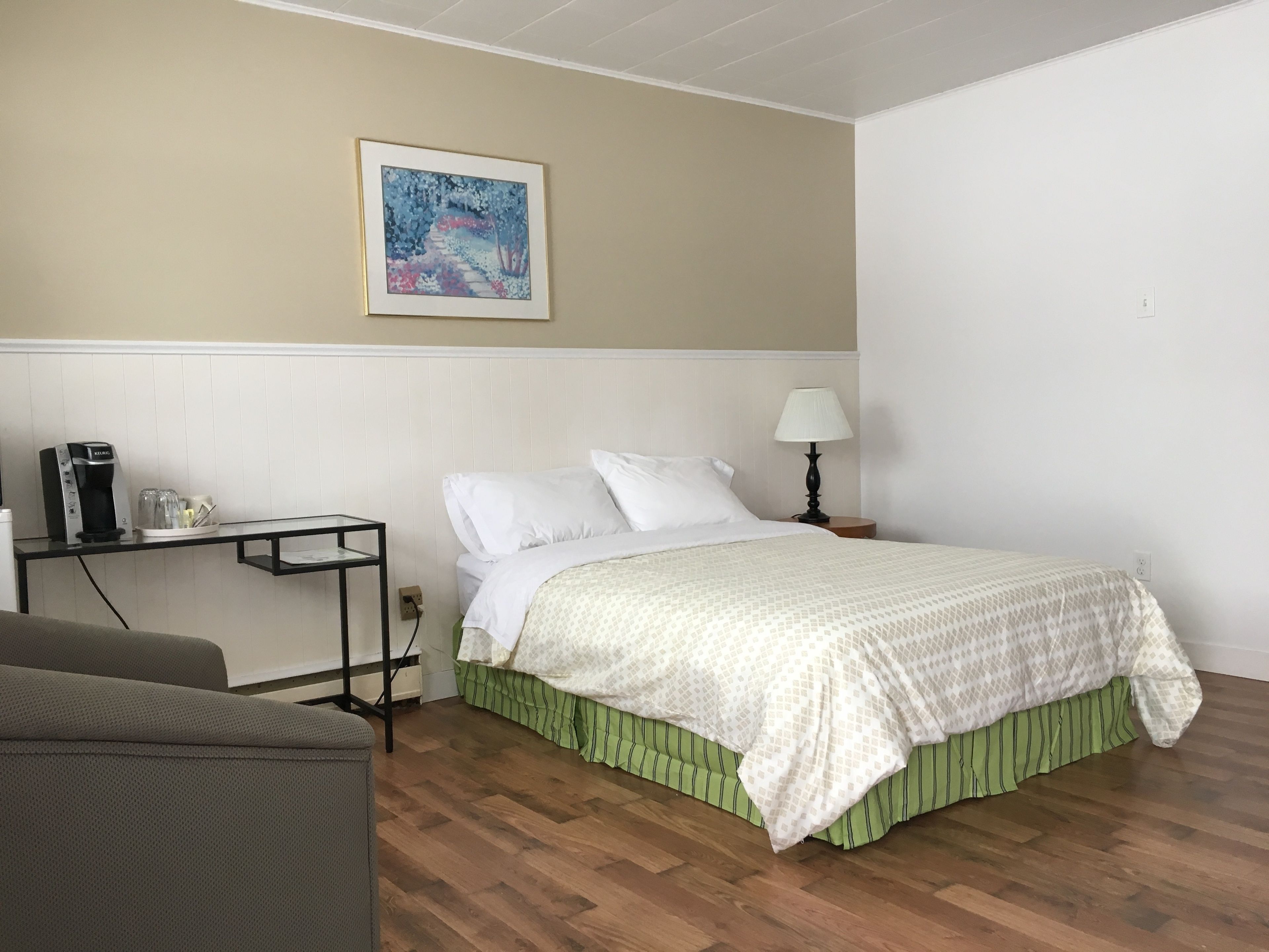 A neatly arranged hotel room with a bed, a bedside table, a lamp, a coffee maker, and a painting on the wall.