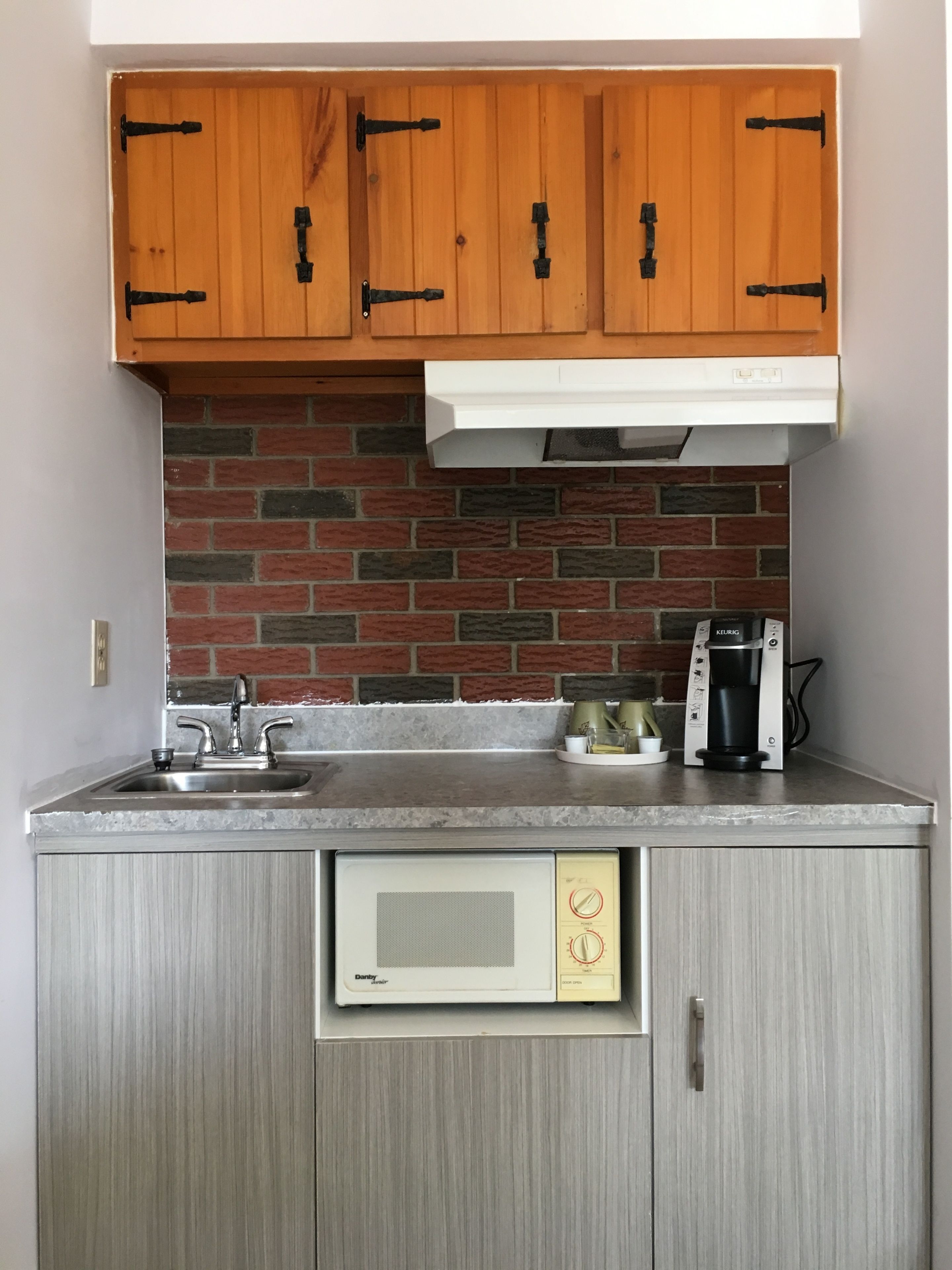 A compact kitchen with wooden cabinets, a brick backsplash, a built-in microwave, and a coffee maker.