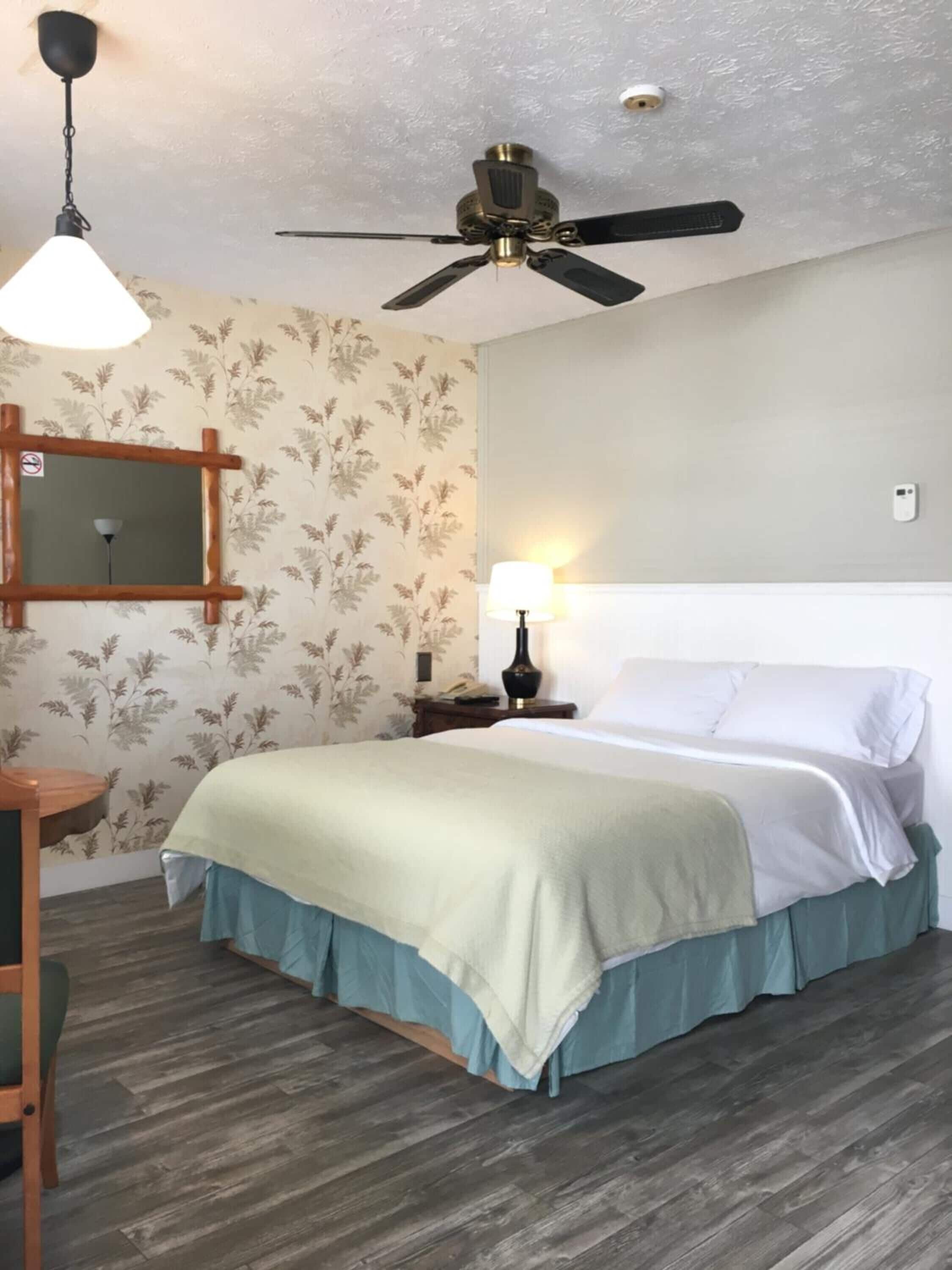 A bedroom with a bed, a nightstand, a lamp, a ceiling fan, a mirror, and a wooden chair.