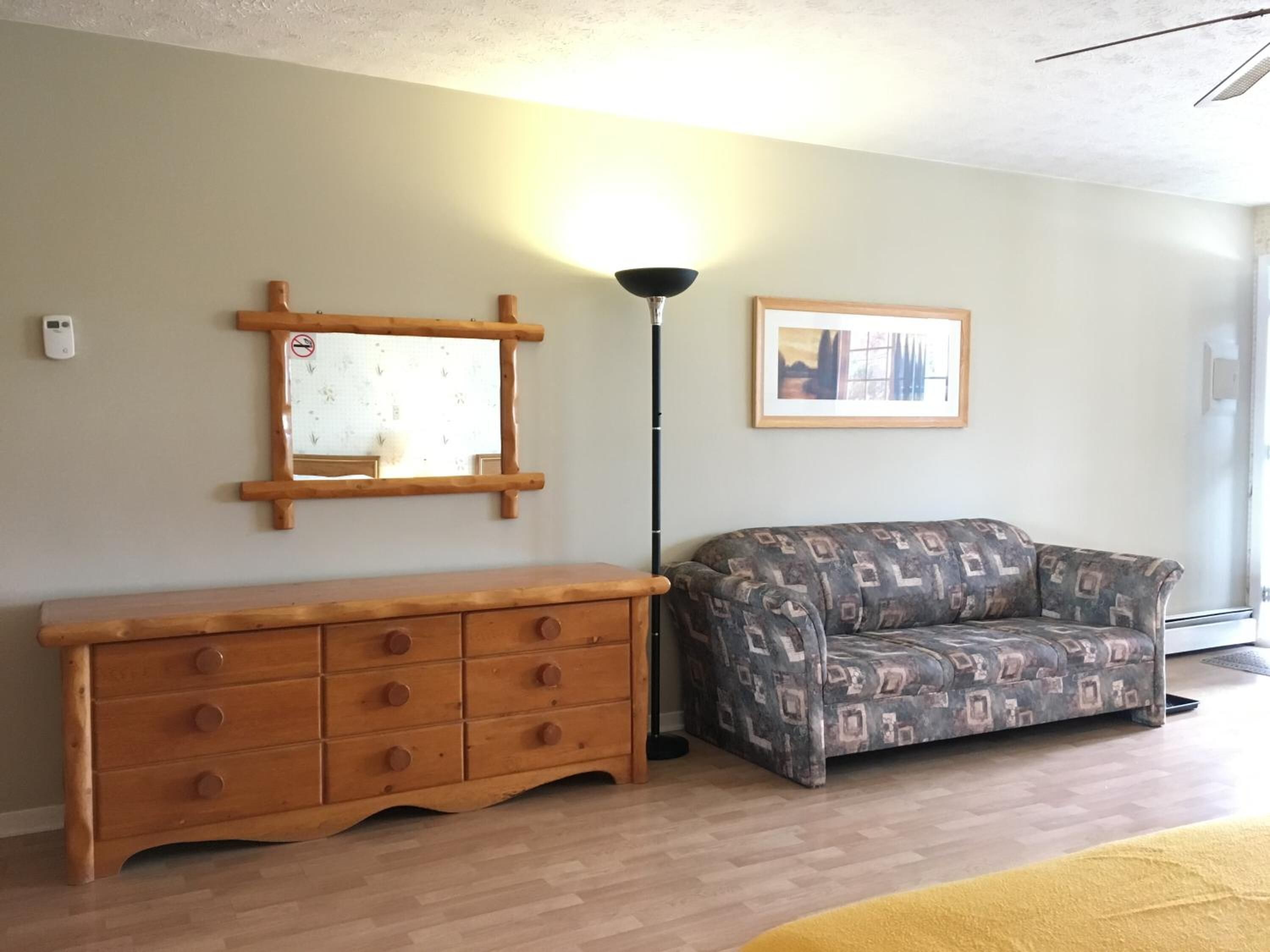 Ein Zimmer mit einem Holzkoffer, einem gemusterten Sofa, einer Stehlampe und einem gerahmten Bild an der Wand.