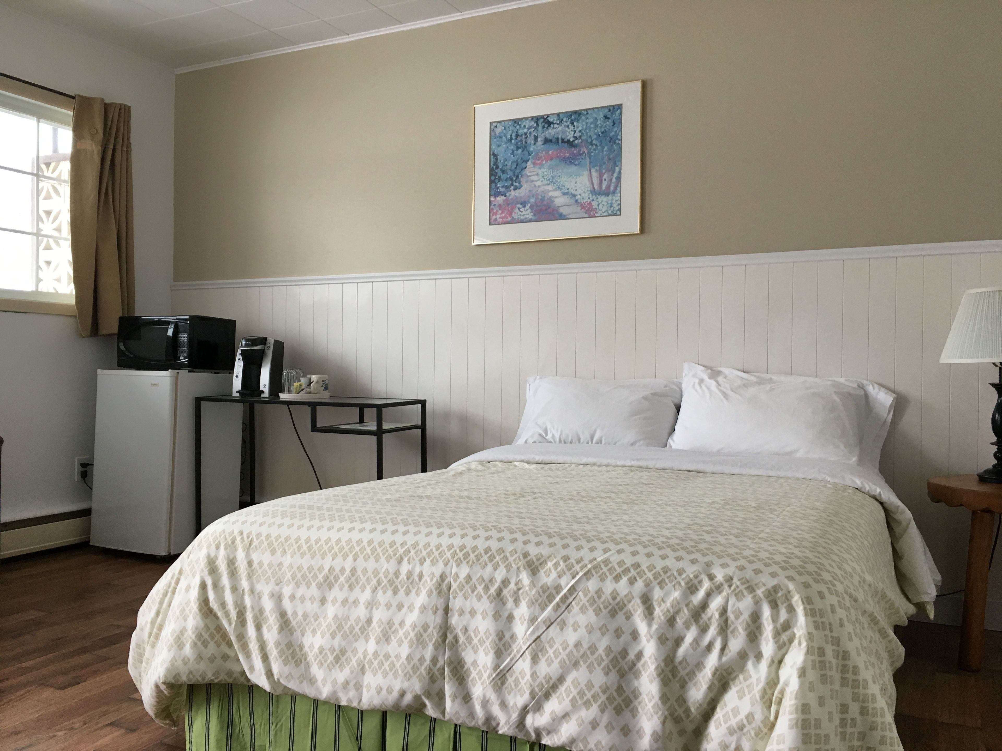 A bedroom with a bed, a nightstand, a desk with a microwave, a refrigerator, a window with curtains, and a painting on the wall.