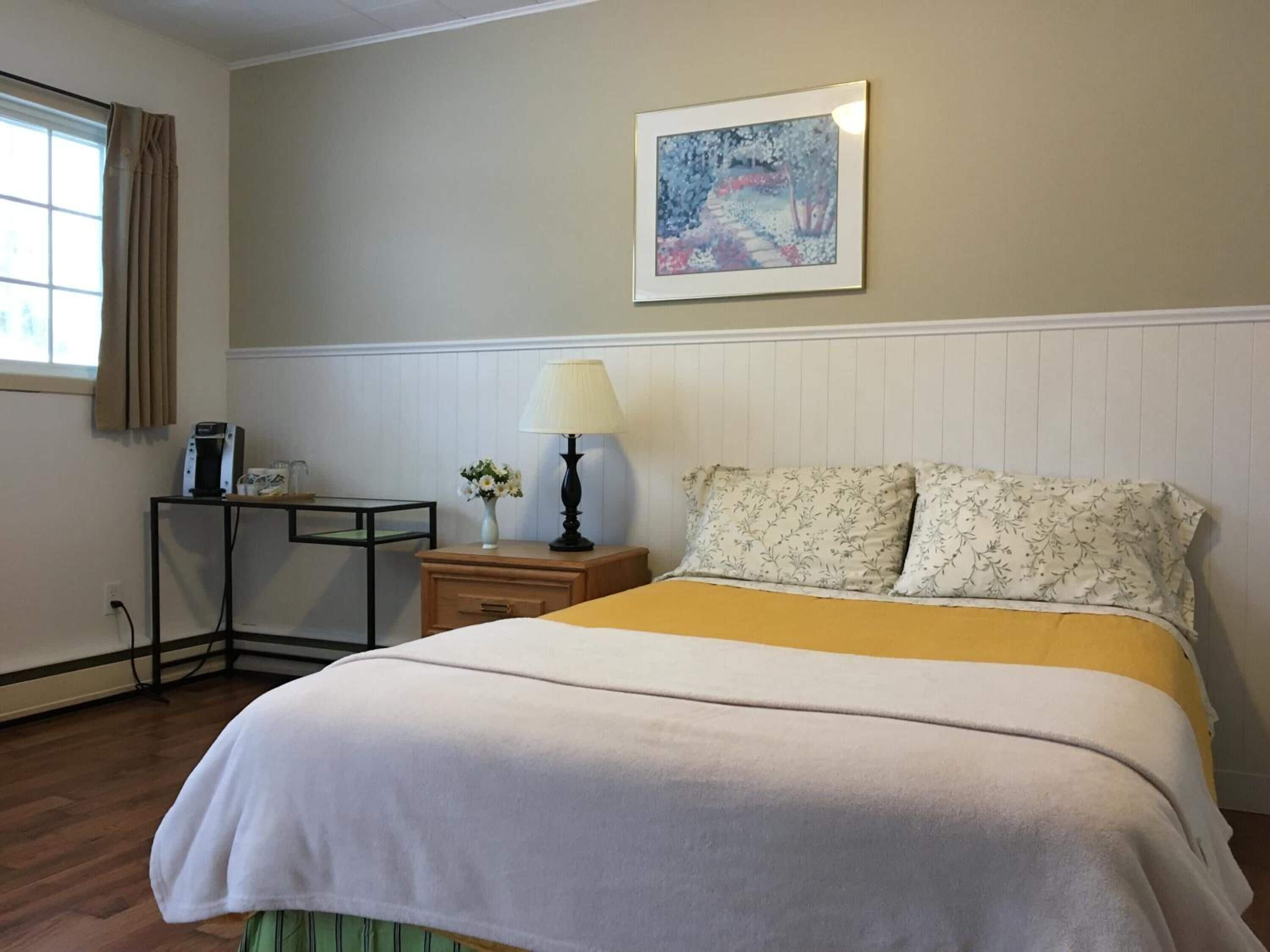 A neatly arranged hotel room with a bed, a bedside table with a lamp, a small table with a coffee maker, and a framed picture on the wall.