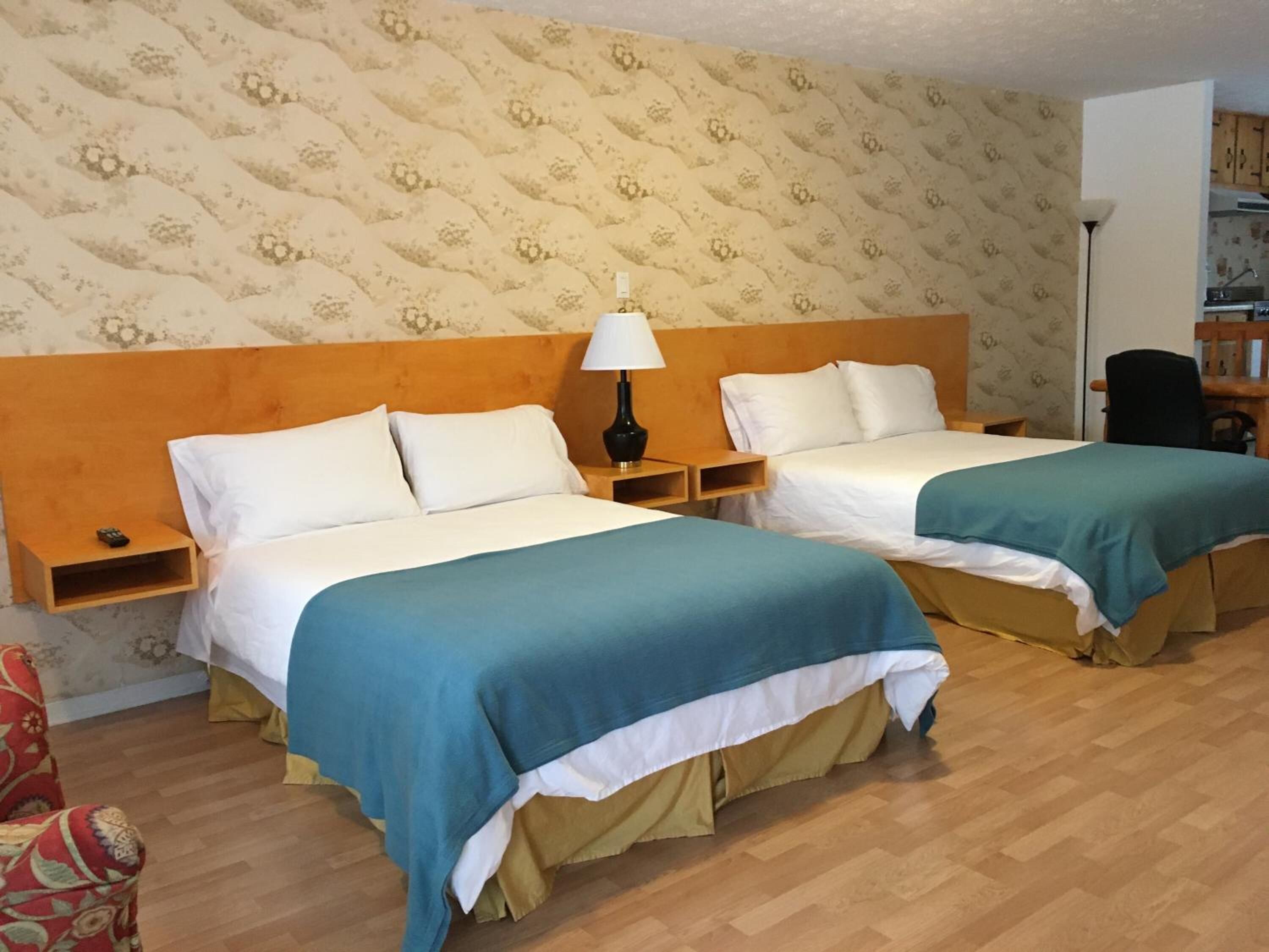 Habitación de hotel con dos camas, cabeceros de madera, una mesita de noche con lámpara, un sillón y un papel tapiz estampado.