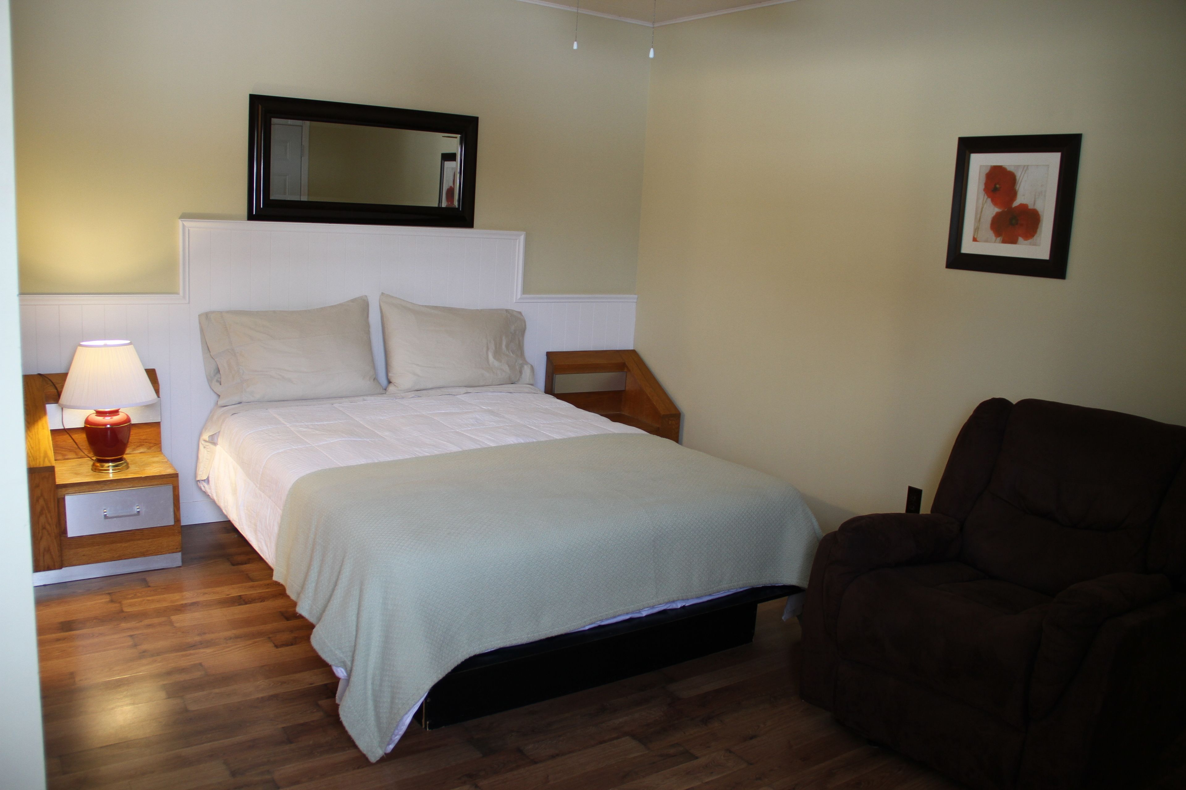 Una camera da letto con un letto, un comodino con una lampada, una sedia, un quadro appeso al muro e uno specchio sopra il letto.