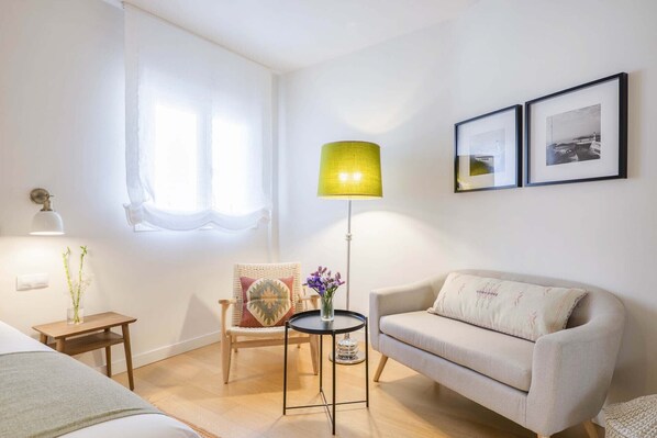 A room with a bed, a chair, a sofa, a small table, a floor lamp, and two framed pictures on the wall.