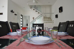 Un coin repas avec une table à plateau de verre, dressée pour quatre personnes, entourée de chaises noires. On aperçoit un escalier à l’arrière-plan.