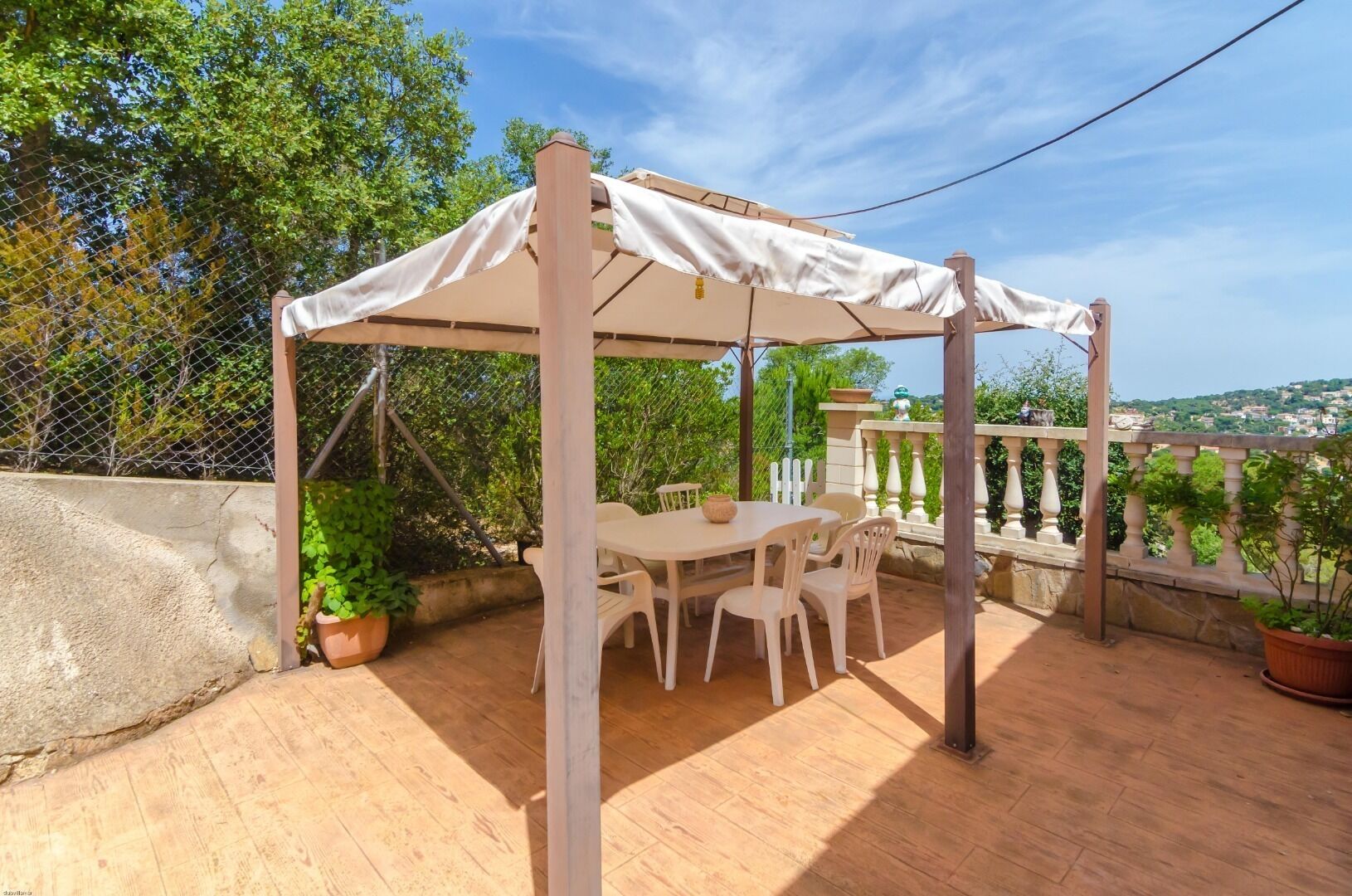 Une terrasse couverte avec une table et des chaises, entourée de plantes en pot et offrant une vue sur la campagne.