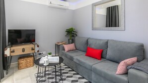 A modern living room with a grey sofa, red and patterned cushions, a flat-screen TV, a small side table, a potted plant, and a decorative mirror. - HAppy House (Cascais)