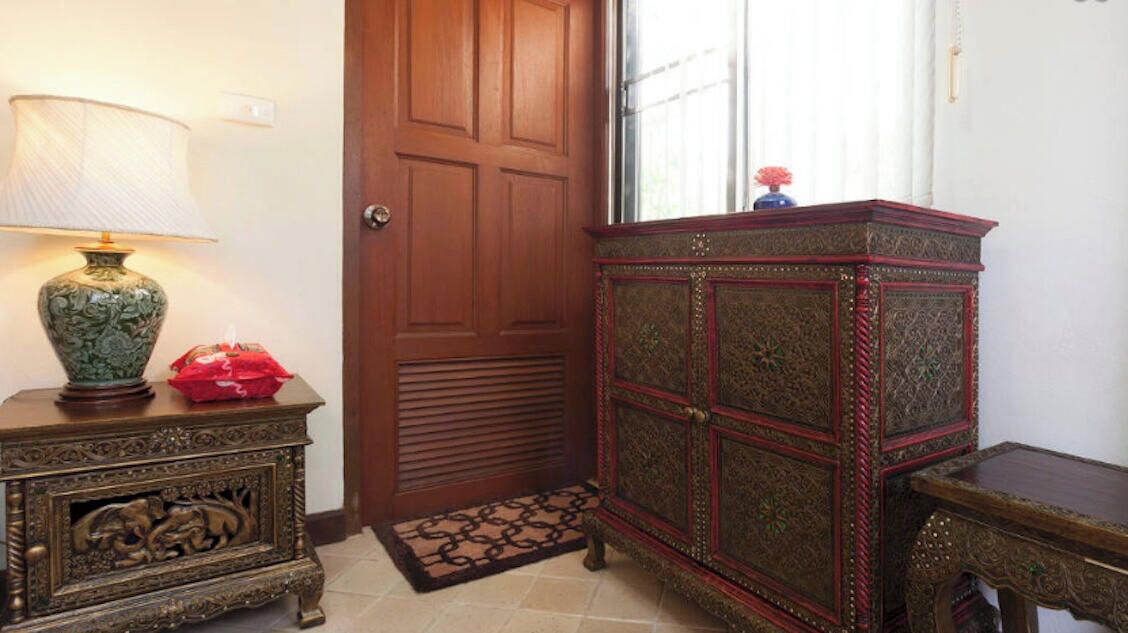 A room with a wooden door, a decorative cabinet, a small table with a lamp, and a red vase.