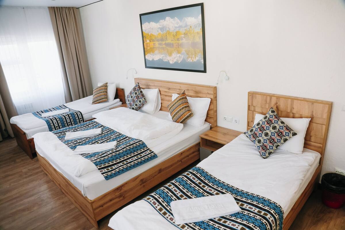 Une chambre d’hôtel avec trois lits en bois, une tête de lit en bois, un tableau encadré au mur et une fenêtre avec des rideaux.