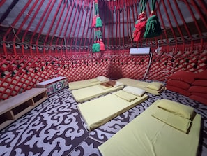 A yurt interior with a patterned floor, multiple beds with pillows, a wooden bench, and a wall-mounted TV.