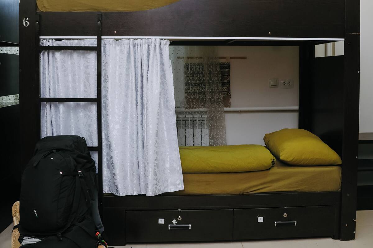 Un lit superposé avec un matelas et des oreillers, une échelle, un sac à dos noir et une fenêtre avec des rideaux.