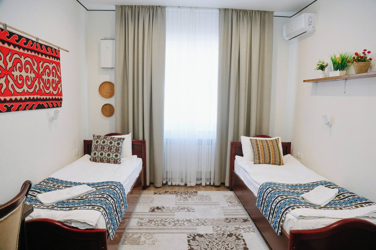 Une chambre d’hôtel avec deux lits simples, une grande fenêtre avec des rideaux, un climatiseur fixé au mur et une étagère avec des objets décoratifs.
