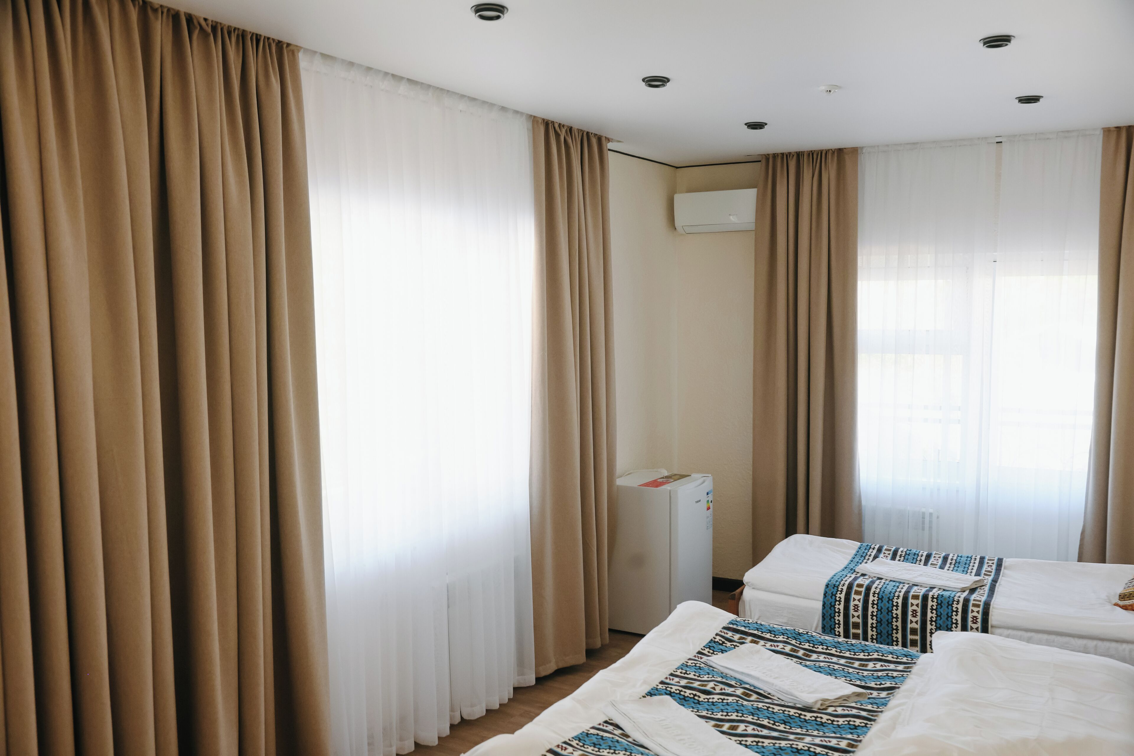 A hotel room with two beds, beige curtains, a white air conditioning unit, and a window with sheer curtains.