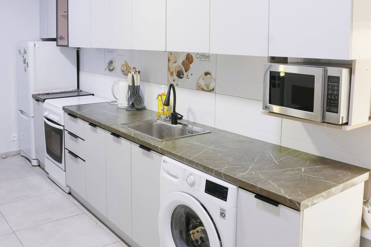 Une cuisine moderne avec des armoires blanches, un four à micro-ondes encastré, un lave-linge et un crédence décorée sur le thème du café.