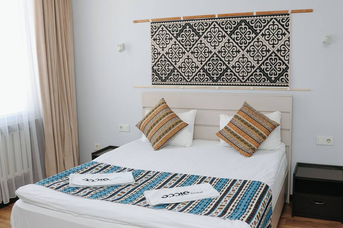 Une chambre d’hôtel comprenant un lit, une tête de lit, une table de chevet, une œuvre d’art accrochée au mur et une fenêtre avec des rideaux.