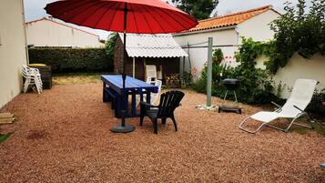 A patio with a table and chairs, a red umbrella, a barbecue grill, and a white lounge chair.