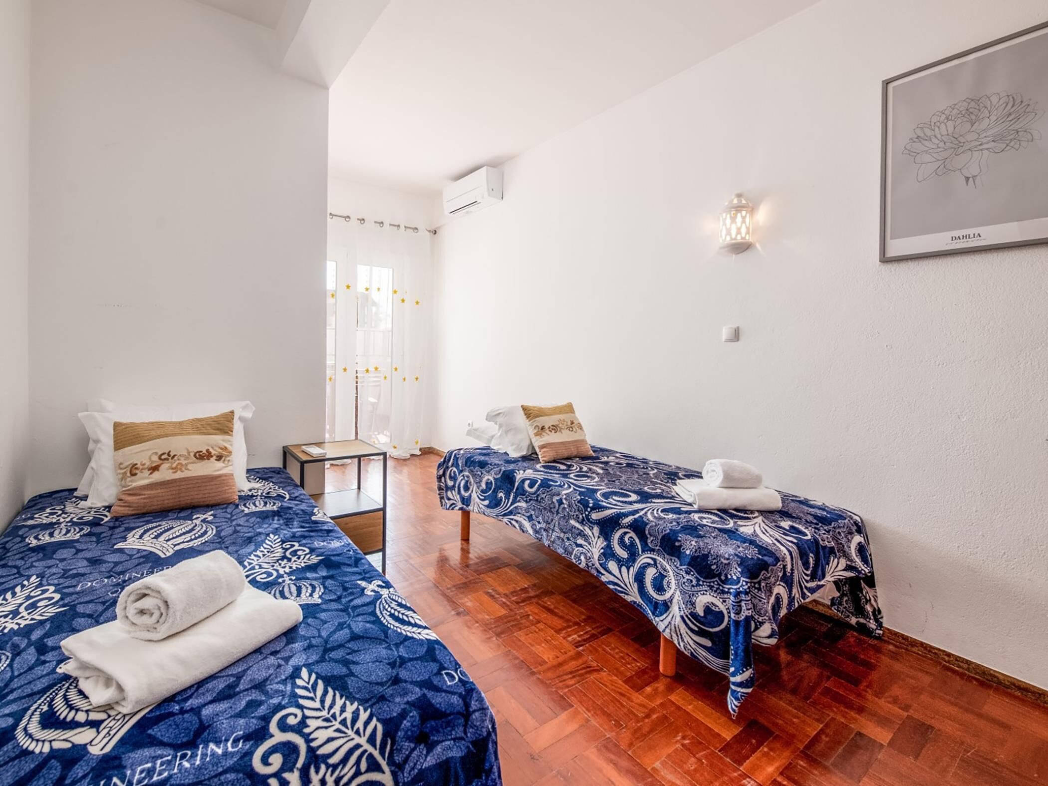 A hotel room with two beds, wooden flooring, a small table, and a window with curtains.