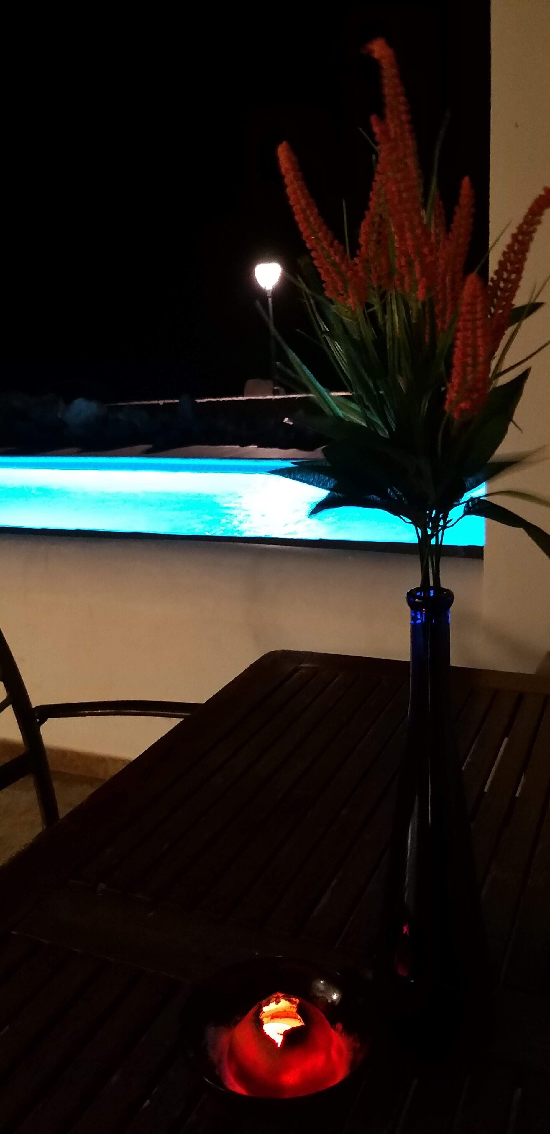 A table with a vase of red flowers and a lit candle, near a pool at night.