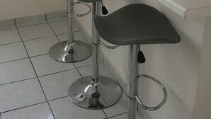 Μια κουζίνα με έναν λευκό πάγκο, τρία σκαμπό μπαρ, ένα παράθυρο με λεπτά, διάφανα κουρτίνια και έ ναν νεροχύτη.