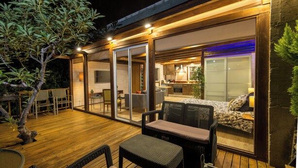 Een terras buiten met houten vloer, rieten meubilair en uitzicht op een modern interieur met keuken en woonruimte.