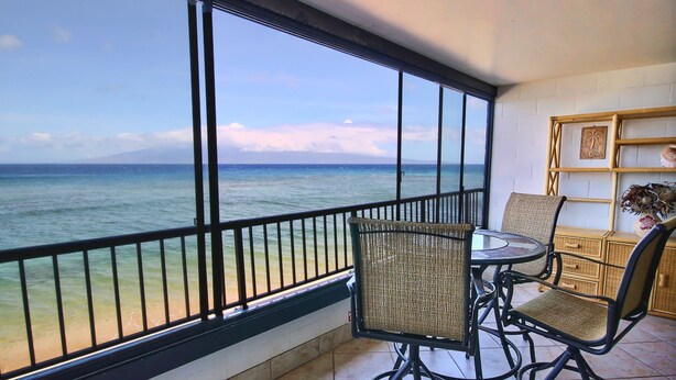 A balcony with a view of the ocean, wicker chairs, a small table, and a decorative shelf with a 'ALOHA' sign.