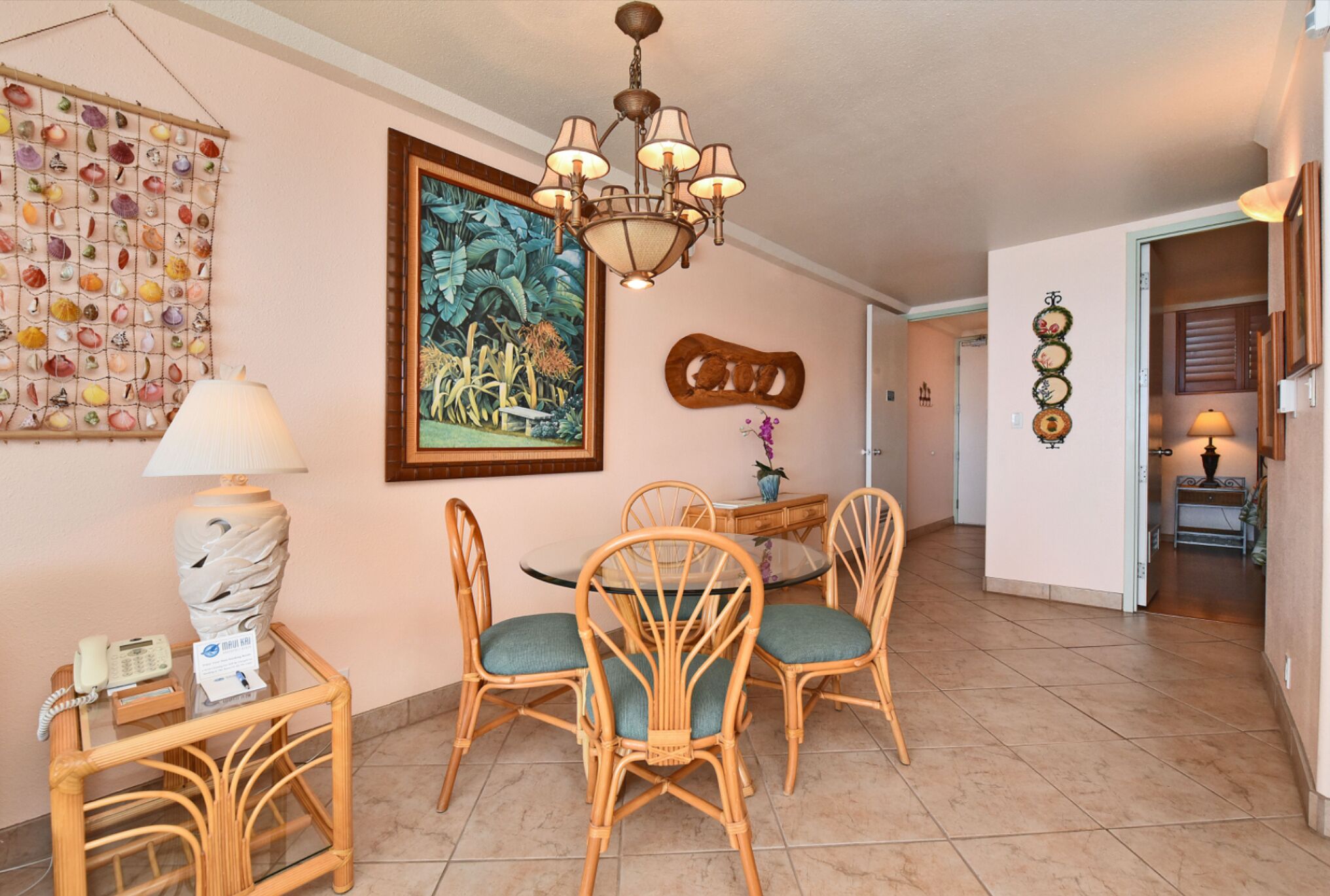 A dining area with wicker chairs and a glass table, a chandelier, a wall art piece, and a telephone on a side table.