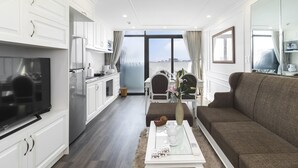 A modern living room with a sofa, a coffee table, and a TV. There is a kitchen area with white cabinets and a refrigerator. The room has a large window with a view of a bridge and buildings.