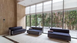 Um lobby moderno com três sofás escuros, piso de mármore e grandes janelas com vista para o verde lá fora.