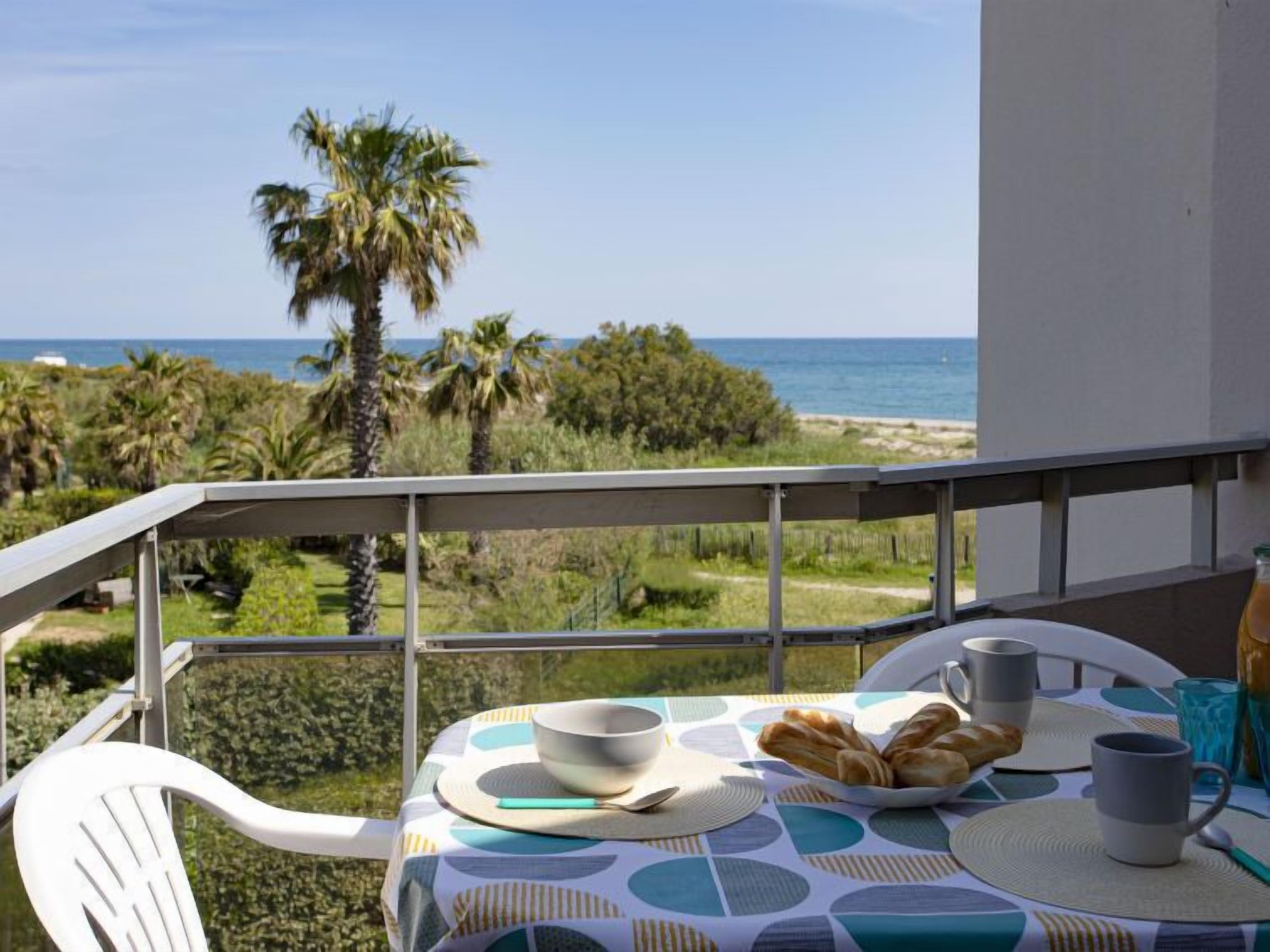 Un balcon avec une table préparée pour le petit-déjeuner, donnant sur une plage et des palmiers.