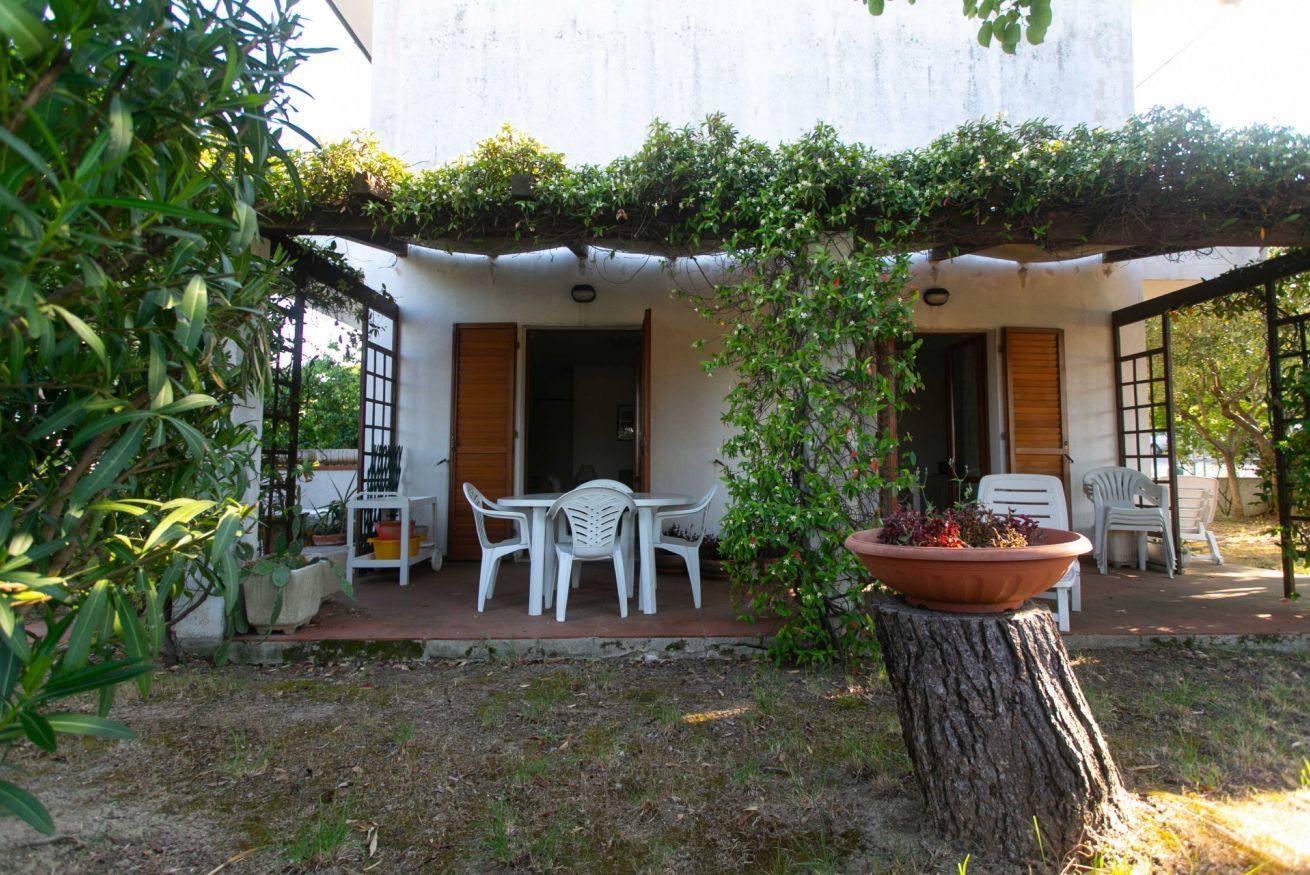 A covered outdoor seating area with white plastic chairs and a table, surrounded by greenery and a tree stump with a potted plant.