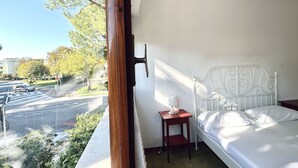 Een balkon met een bed, een kleine tafel met een lamp en uitzicht op een straat en gebouwen.