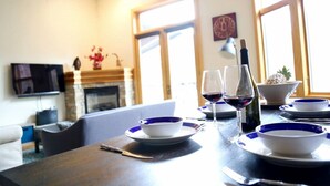 Una mesa de comedor dispuesta con platos blancos y azules, dos copas de vino y una botella de vino. Una televisión está montada en la pared, y se observa una chimenea al fondo.