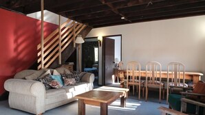 Uma sala de estar com sofá, mesa de centro e cadeiras de madeira. Há uma escada com uma parede em destaque vermelha e uma porta que dá para outro cômodo.