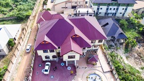 A multi-story building with a distinctive red roof, a driveway with parked cars, and a small garden area. - Gya-son Royal Guesthouse (Kumasi)
