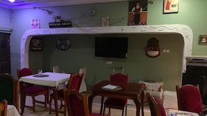 Un espace repas avec des tables en bois et des chaises à revêtement rouge, un mur décoratif blanc et un téléviseur fixé au-dessus d’une table.