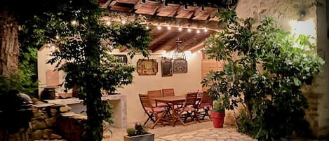 Un patio al aire libre con un rústico muro de piedra, mobiliario de madera y luces colgantes.