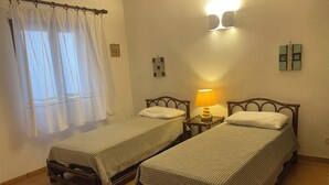 A room with two wooden beds, a small table with a lamp, a window with sheer curtains, and a wall-mounted light fixture.