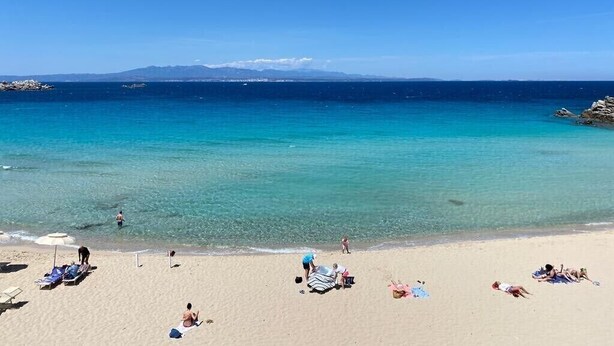A beach with clear turquoise water, people sunbathing, and a few beach chairs.