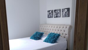 A bedroom with a tufted headboard, a white bedspread, and two blue pillows. There are three framed pictures on the wall above the bed. - Gran apartamento en plaza principal (Guatape)
