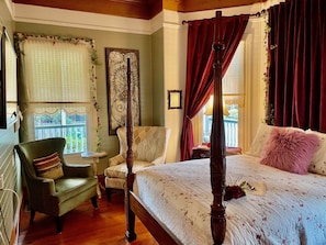 A bedroom with a four-poster bed, a green armchair, a small table, and a window with curtains.