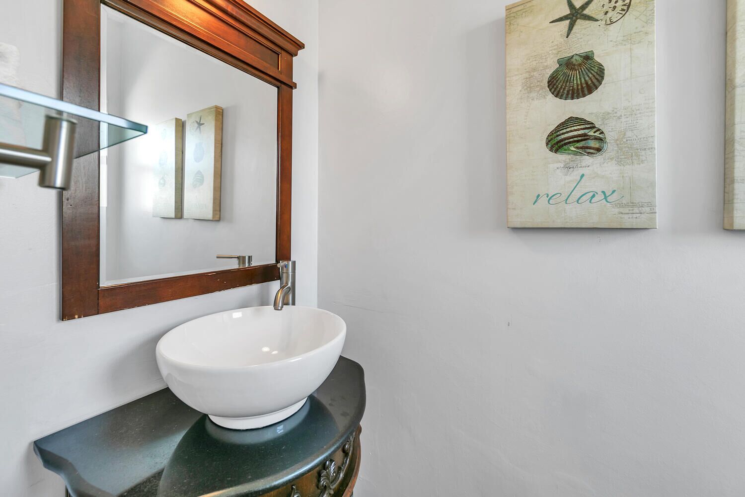 Un baño con un lavamanos blanco tipo pedestal, un espejo con marco de madera y una obra de arte en la pared con conchas marinas y la palabra 'relax'.