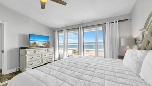 Een slaapkamer met een groot bed, een dressoir, een televisie waarop een strandscène te zien is en uitzicht op het strand door de ramen.
