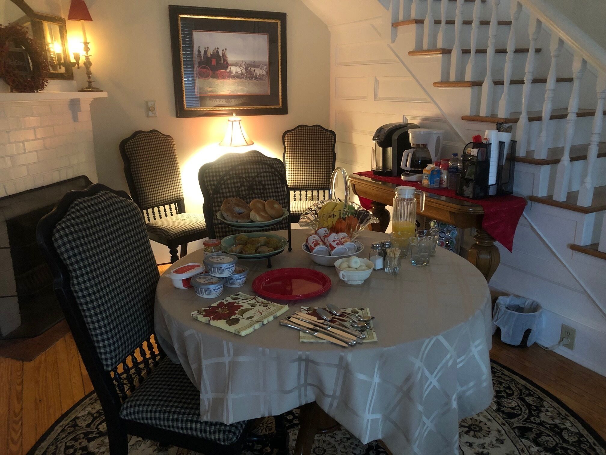 A dining table set with food and drinks, surrounded by checkered chairs, in a room with a staircase and a framed picture on the wall.