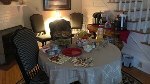 A dining table set with food and drinks, surrounded by checkered chairs, in a room with a staircase and a framed picture on the wall.