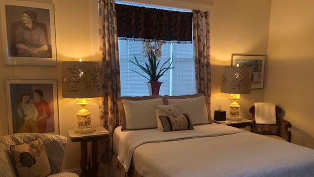 A bedroom with a bed, two nightstands, a chair, a potted plant, and artwork on the walls.